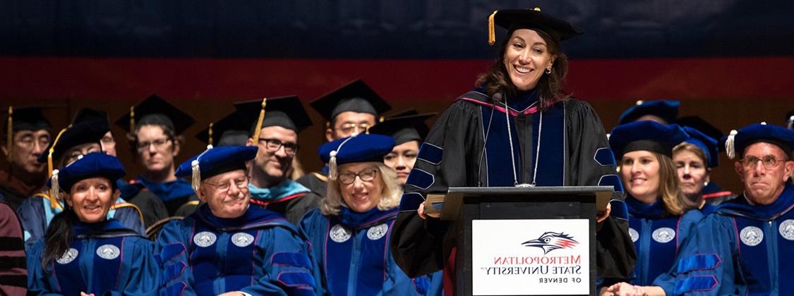 Photograph of President Janine Davidson in her graduation gown at a graduation ceremony with several department chairs sitting behind her in their graduation gowns.